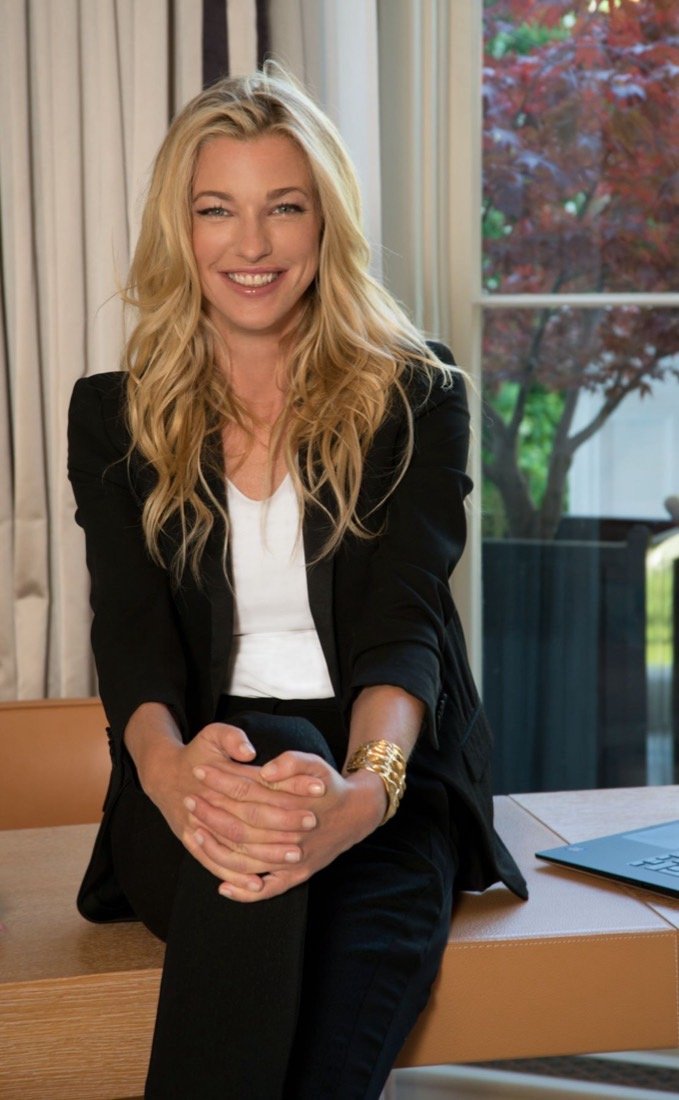 Nicole Junkerman smiling warmly while seated on the edge of a desk beside a bright window.