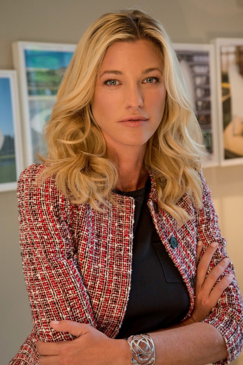 Nicole Junkerman standing with her arms folded in a red and white tweed jacket, framed pictures on the wall behind her.