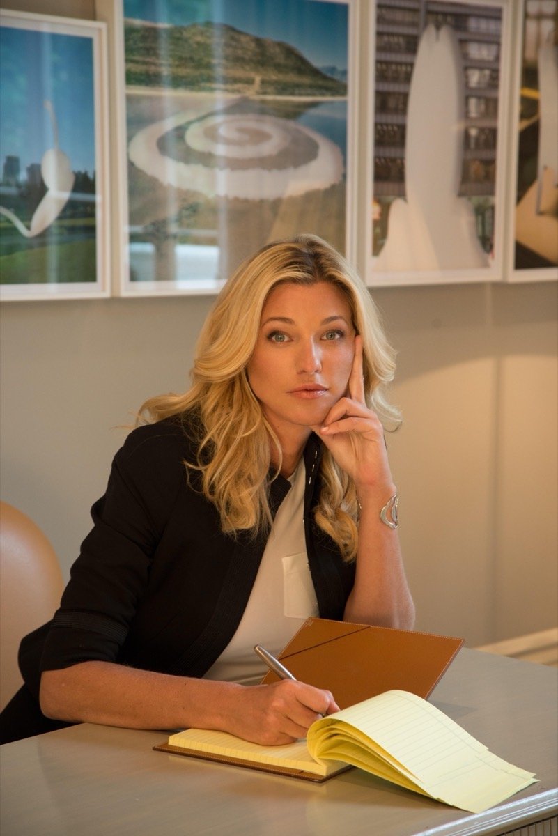 Nicole Junkerman writing thoughtfully in a notebook at a desk with framed prints behind her.